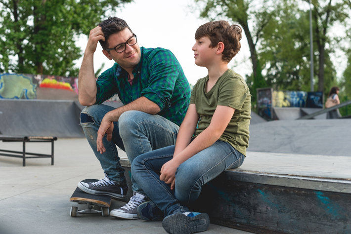 Papa e hijo con una patineta sentados en un parque para patinaje